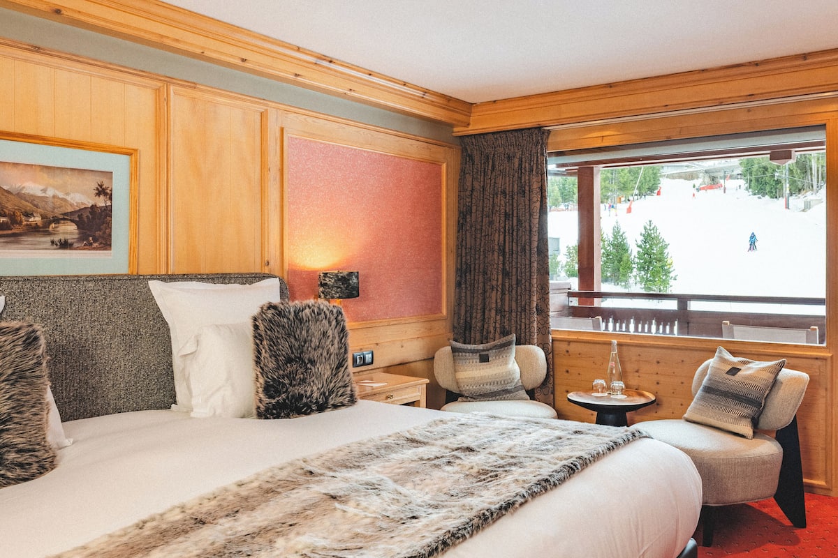 Habitación de hotel con una cama grande, cabecera de madera, ventana con vistas a un paisaje nevado, una mesita con una lámpara y una silla.