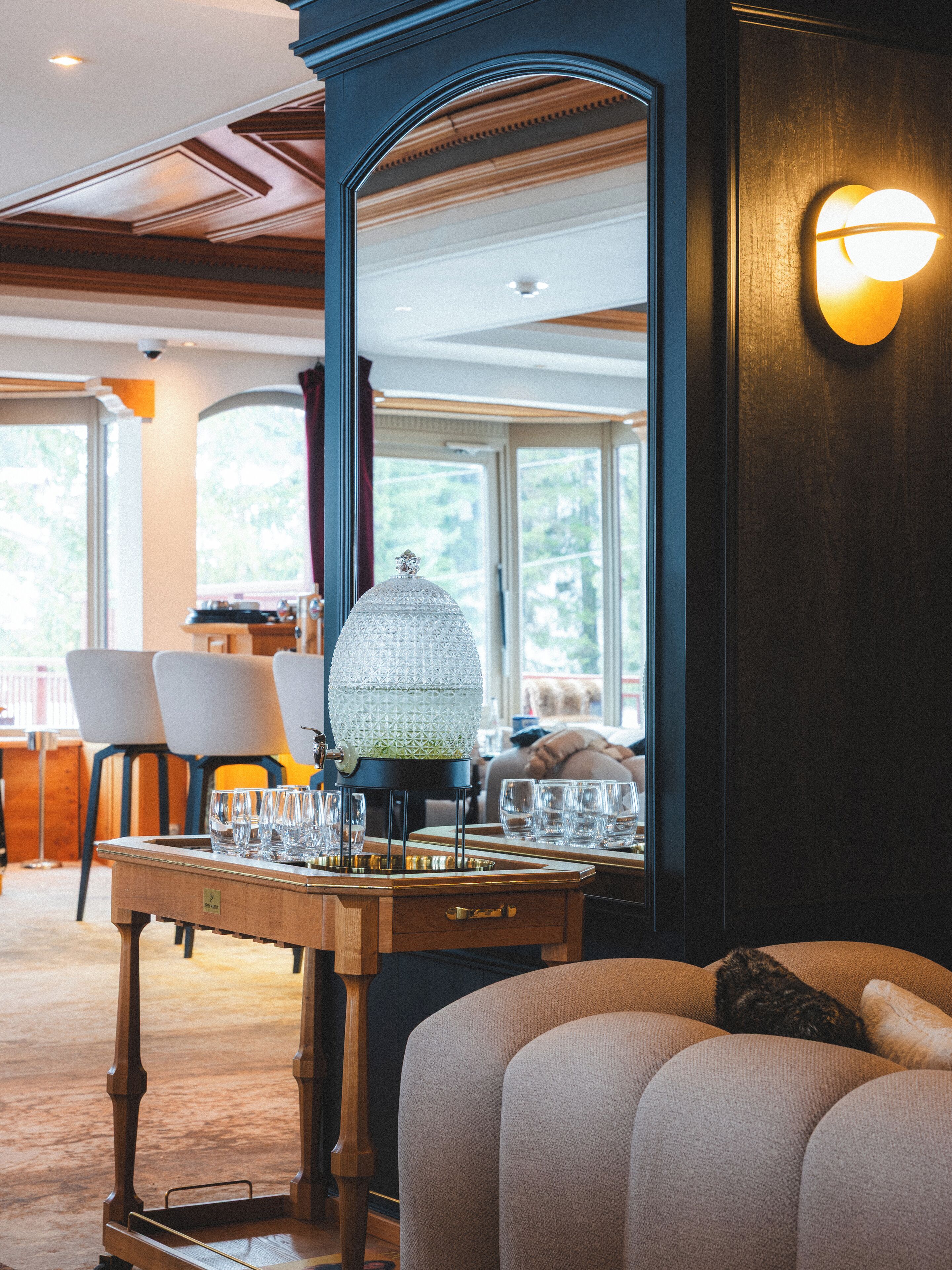 A cozy room with a wooden table set with glasses, a decorative lamp, and a large mirror reflecting the room.