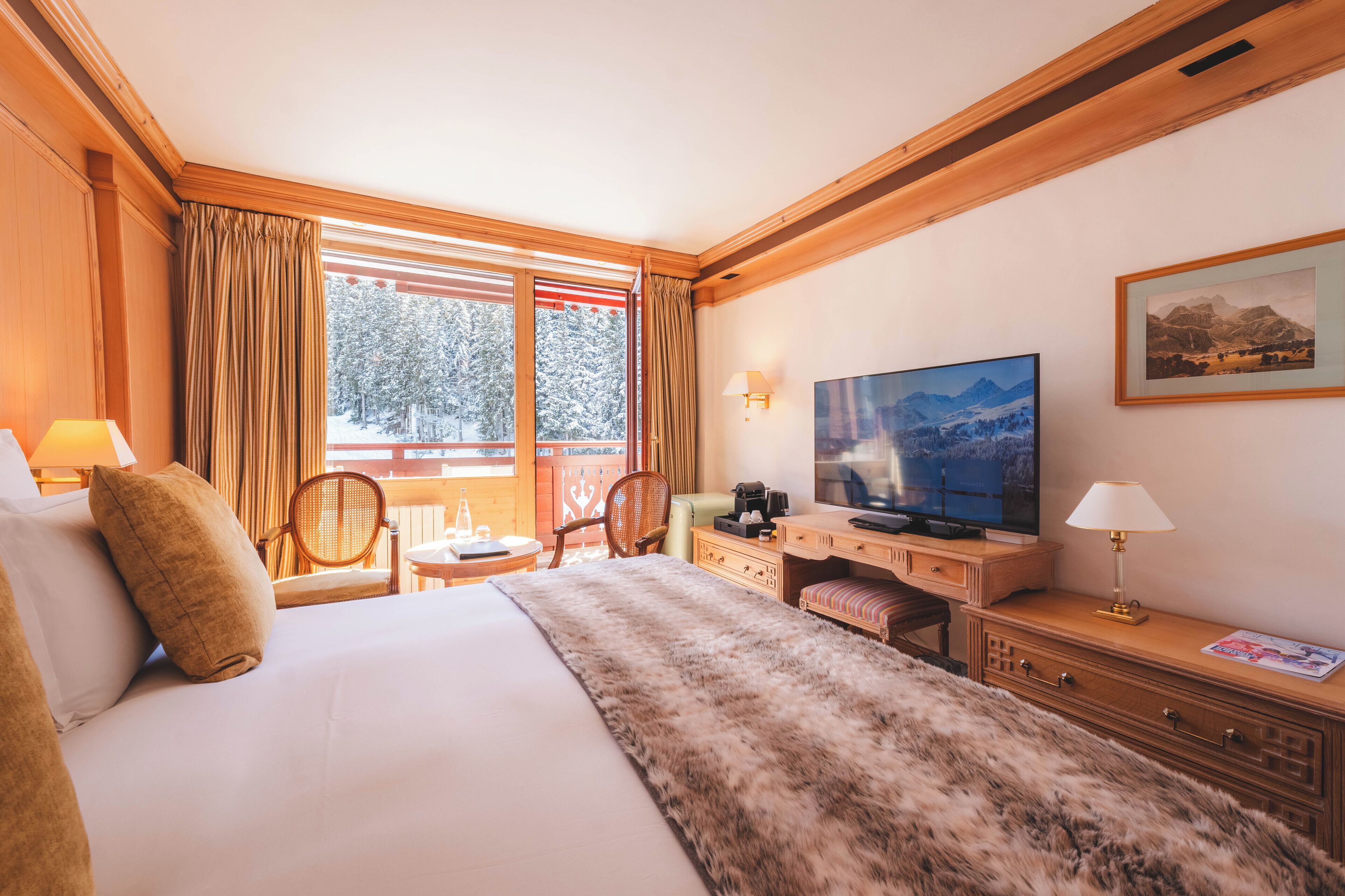 A hotel room with a large bed, a desk with a television, a chair, and a view of snow-covered trees through the window.