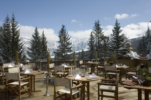 Un área de comedor al aire libre con mesas y sillas de madera, frente a un paisaje de montañas nevadas y árboles de hoja perenne.