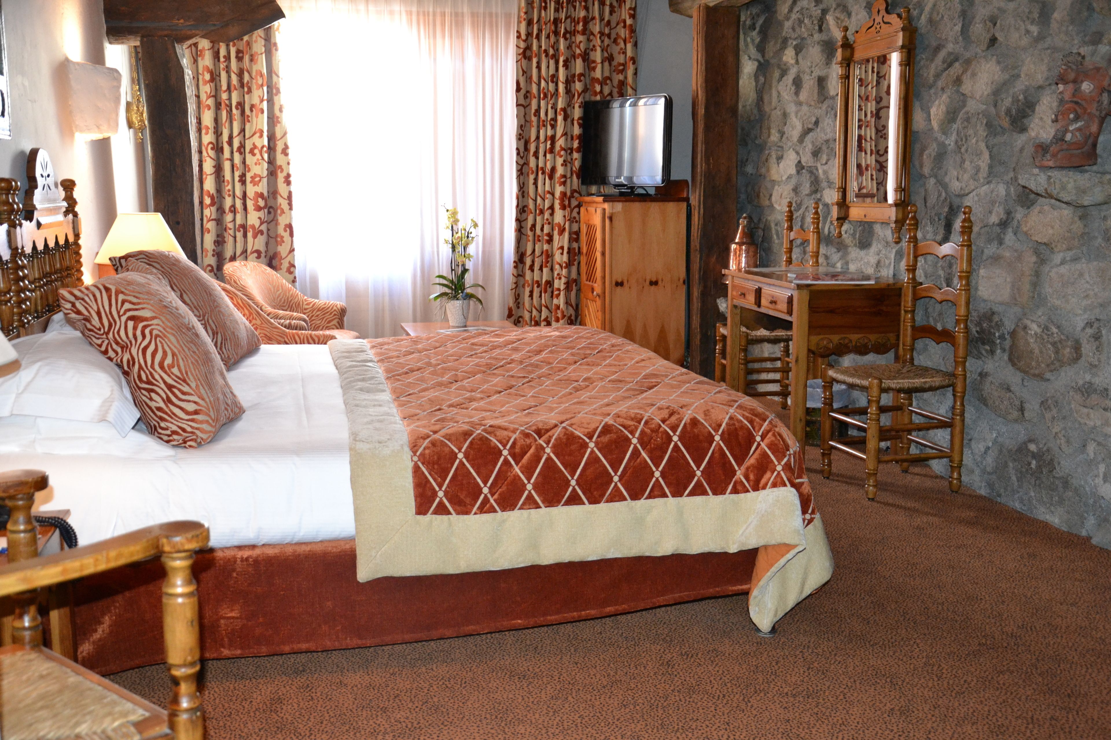 A bedroom with a large bed, a wooden desk, a chair, a television, and a stone wall.