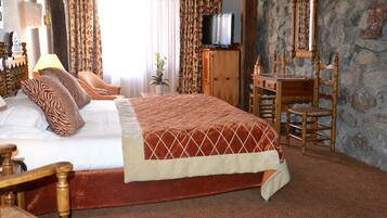 A bedroom with a large bed, a wooden desk, a chair, a television, and a stone wall.
