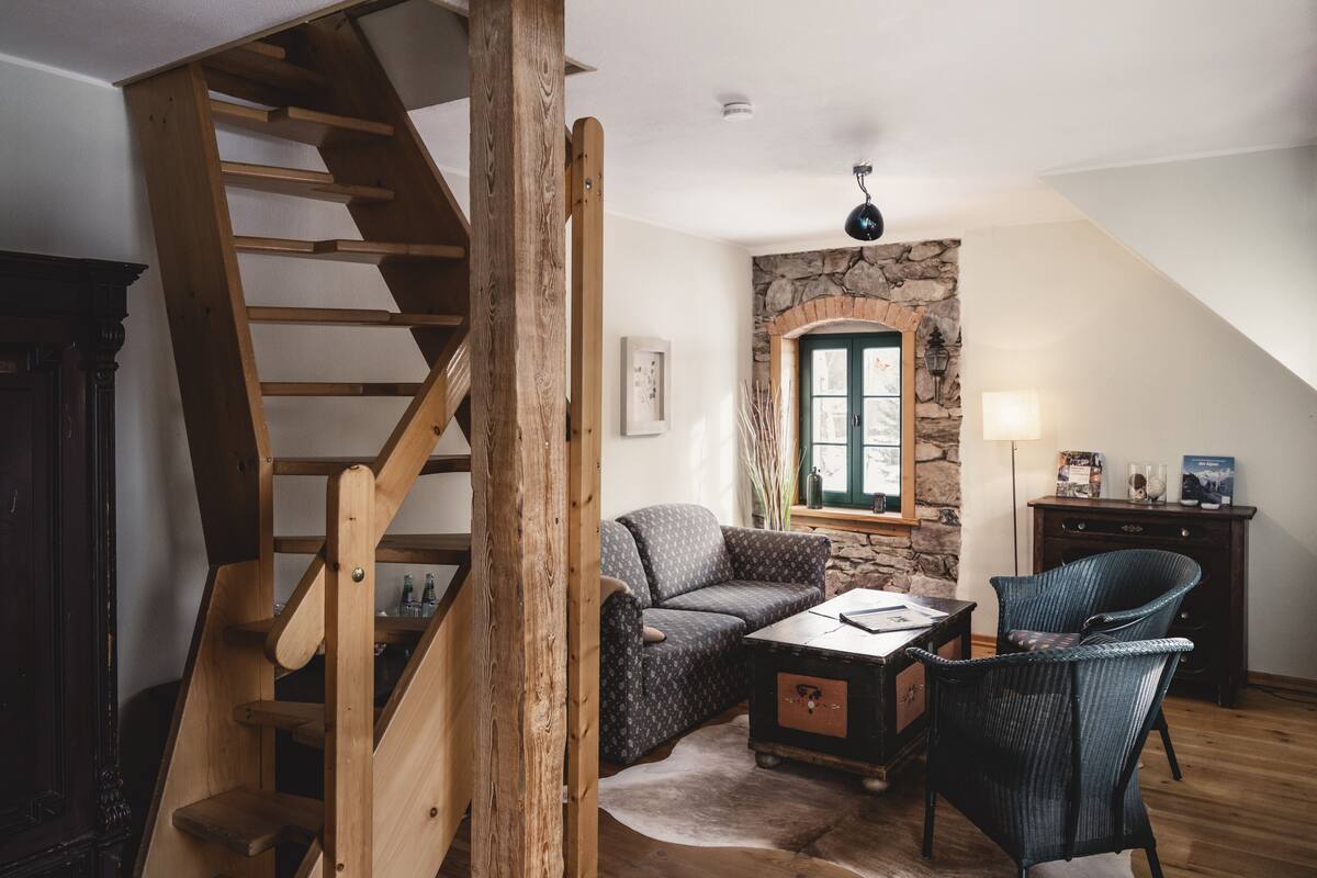Une pièce chaleureuse avec un escalier en bois, un mur en pierre, une porte verte, un canapé, une table basse et un fauteuil.