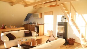 A cozy living room with a sofa, coffee table, and a fireplace. There is a kitchen area with cabinets and a stove. A wooden staircase is visible in the background. - Altsächsischer Gasthof Kleines Vorwerk (Sayda)