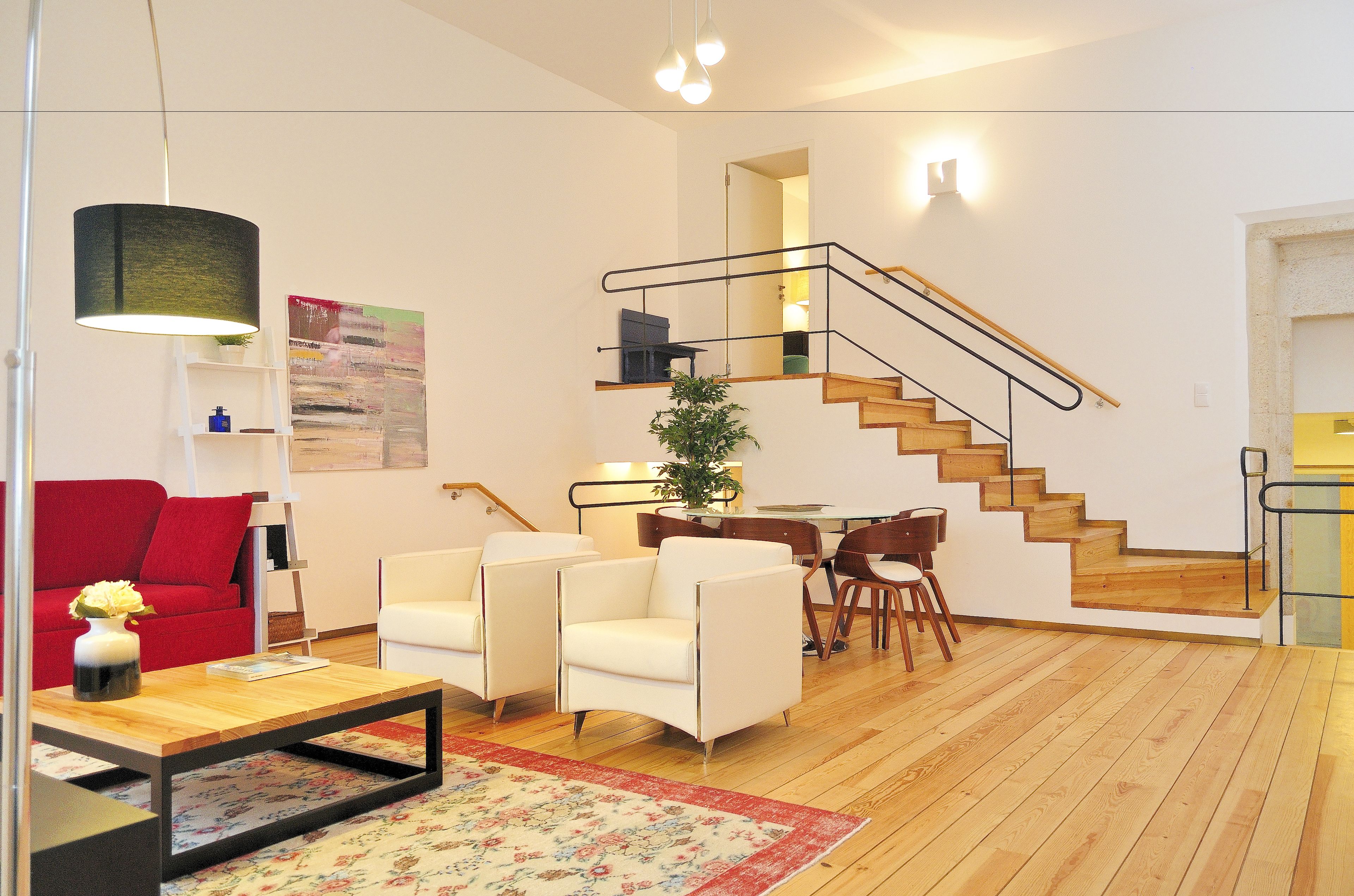 Ein modernes Wohnzimmer mit einem roten Sofa, weißen Sesseln, einem Holztisch und einer Treppe, die zu einer oberen Ebene führt.