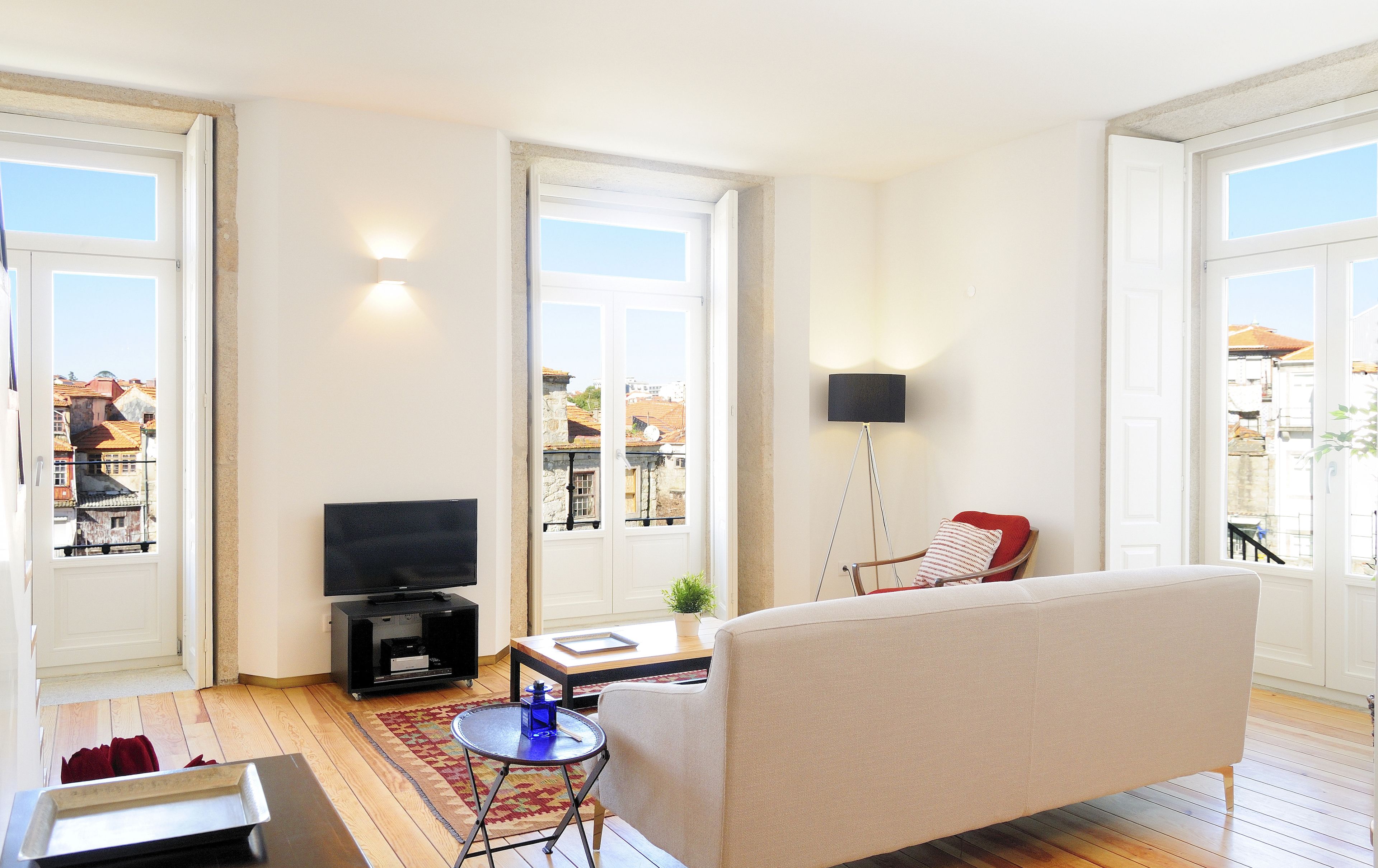 A living room with a sofa, a TV, a small table, and a chair. There are windows with views of buildings outside.