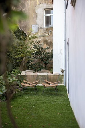 Ein kleiner, abgeschlossener Außenbereich mit zwei Korbstühlen und einem Betontisch, umgeben von einer weißen Mauer und einem Garten mit verschiedenen Pflanzen.