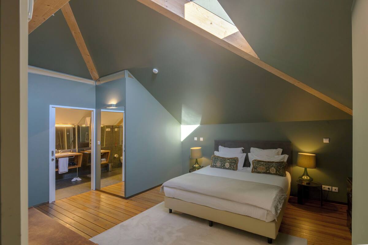 Une chambre à coucher avec un grand lit, un plancher en bois, une lucarne et une salle de bain visible à travers une porte ouverte.