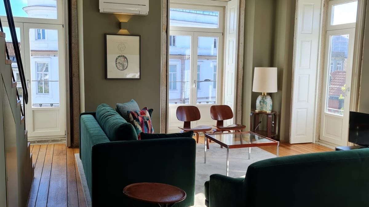 A living room with a green sofa, two armchairs, a coffee table, and a TV. There are two windows with curtains, a framed picture on the wall, and a lamp on a side table.