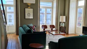 A living room with a green sofa, two armchairs, a coffee table, and a TV. There are two windows with curtains, a framed picture on the wall, and a lamp on a side table.