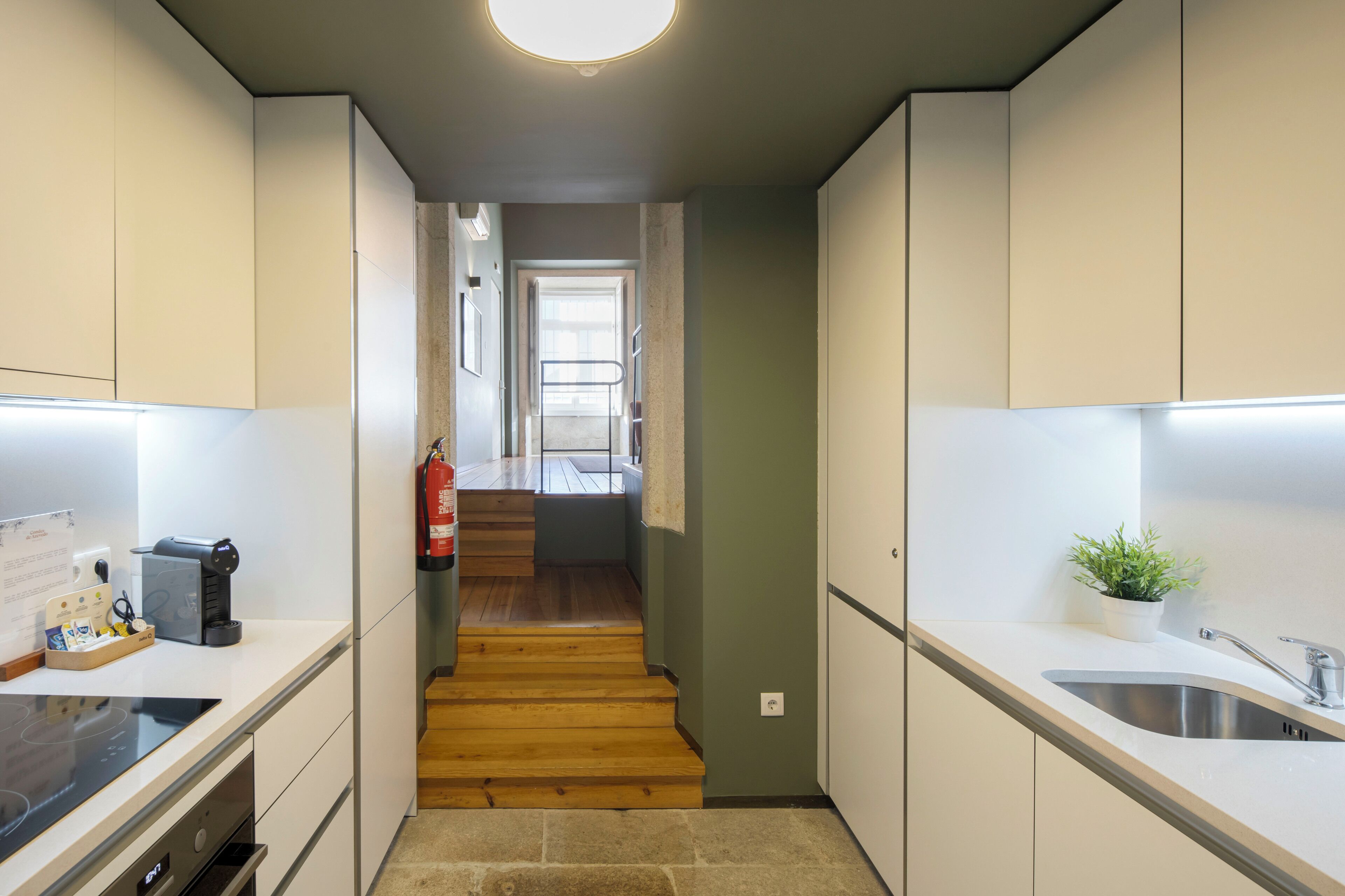 A narrow kitchen with wooden flooring, white cabinets, a stainless steel sink, and a small potted plant.