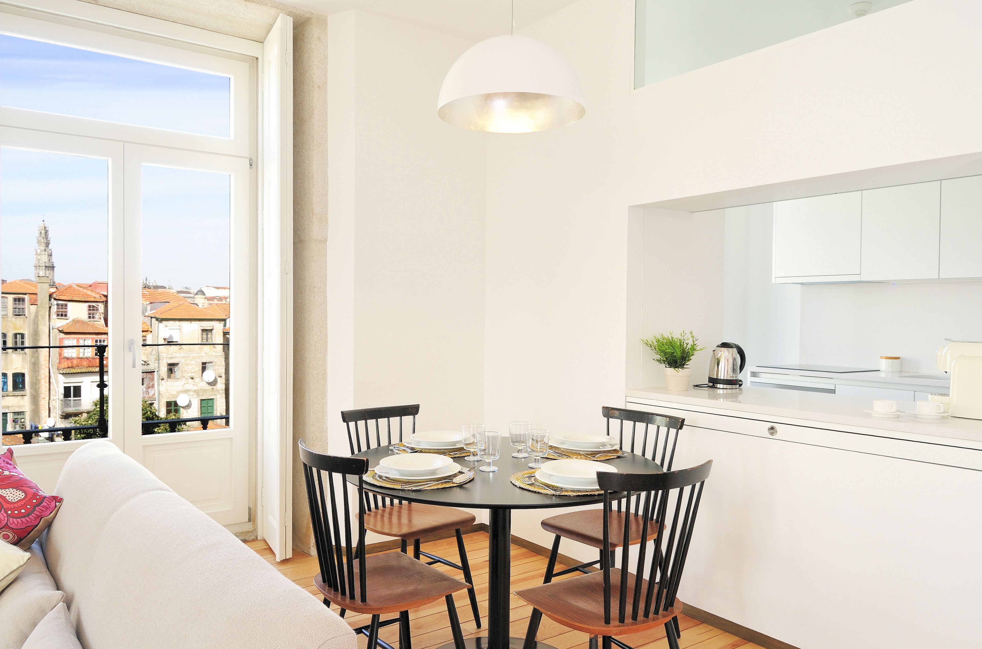 Un coin repas avec une table ronde dressée pour quatre personnes, un canapé avec un coussin et une vue sur les bâtiments par une fenêtre.