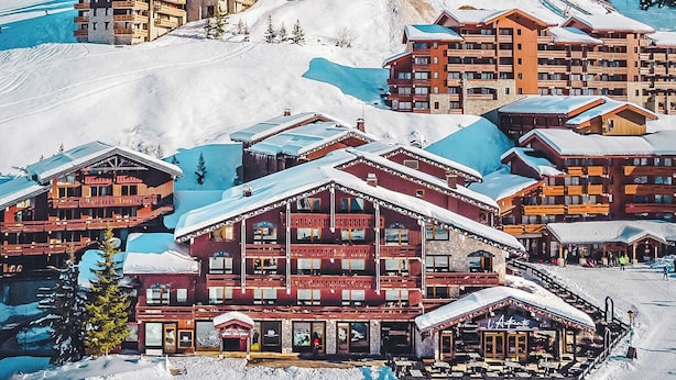 Een bergresort bedekt met sneeuw, met meerdere houten chalets, skipistes en een helderblauwe lucht.