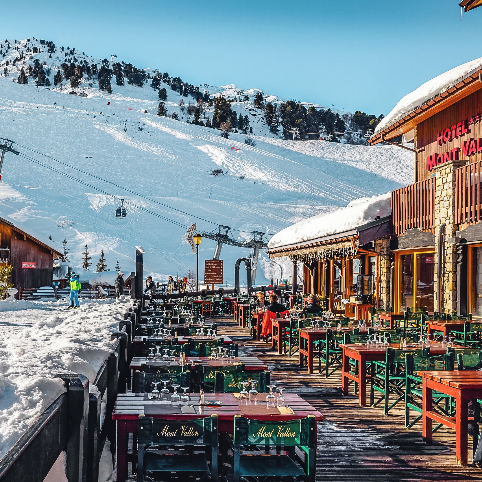 A snow-covered outdoor dining area with tables and chairs, a wooden building with a sign reading "Hotel Mont Vallon", and a ski lift in the background.