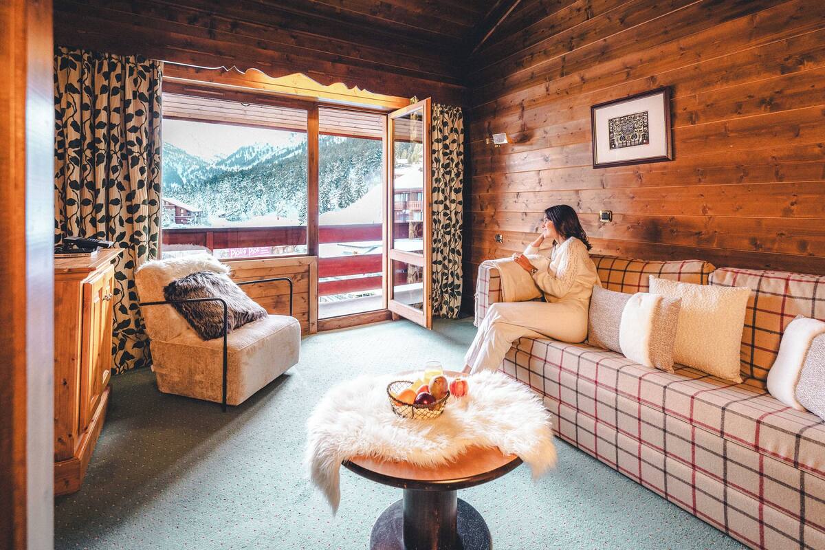 A cozy cabin interior with a sofa, a chair, a table with fruit, and a view of mountains through a large window.