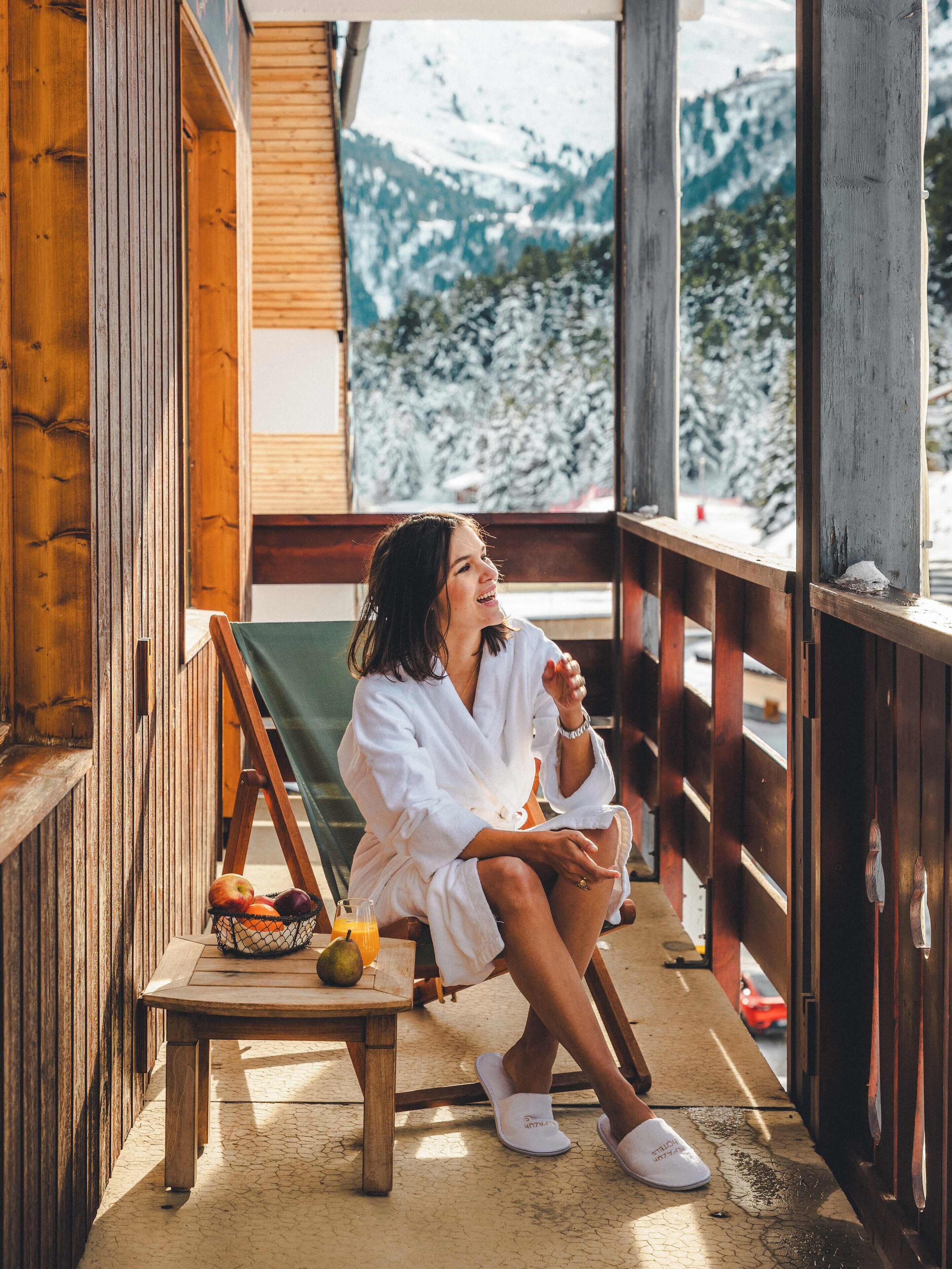 A person seated on a wooden bench in a wooden cabin with a view of snow-covered mountains.