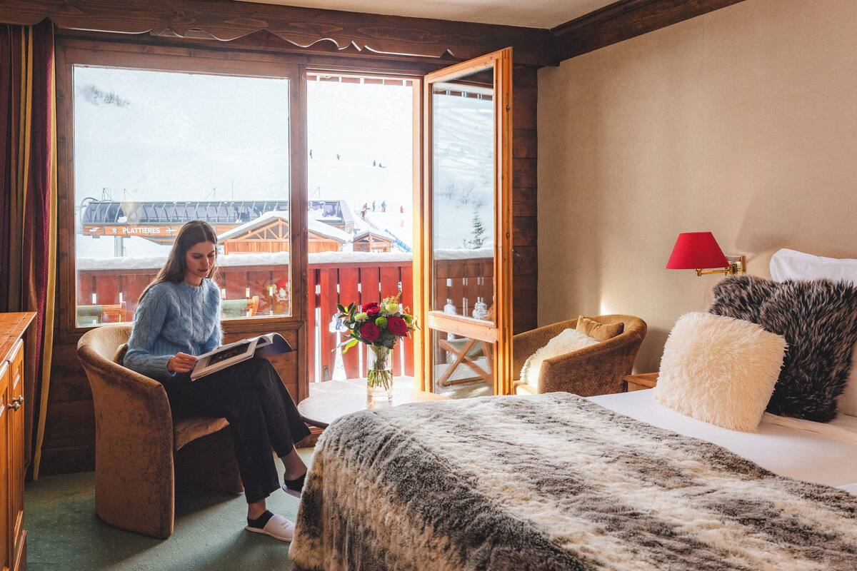 A hotel room with a bed, a chair, a desk, and a window with a view of buildings and a snowy landscape.
