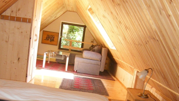 An attic bedroom with a bed, a small table, a lamp, and a window with a view of greenery. - Ferienhaus Leitenhof (Döbrököz)