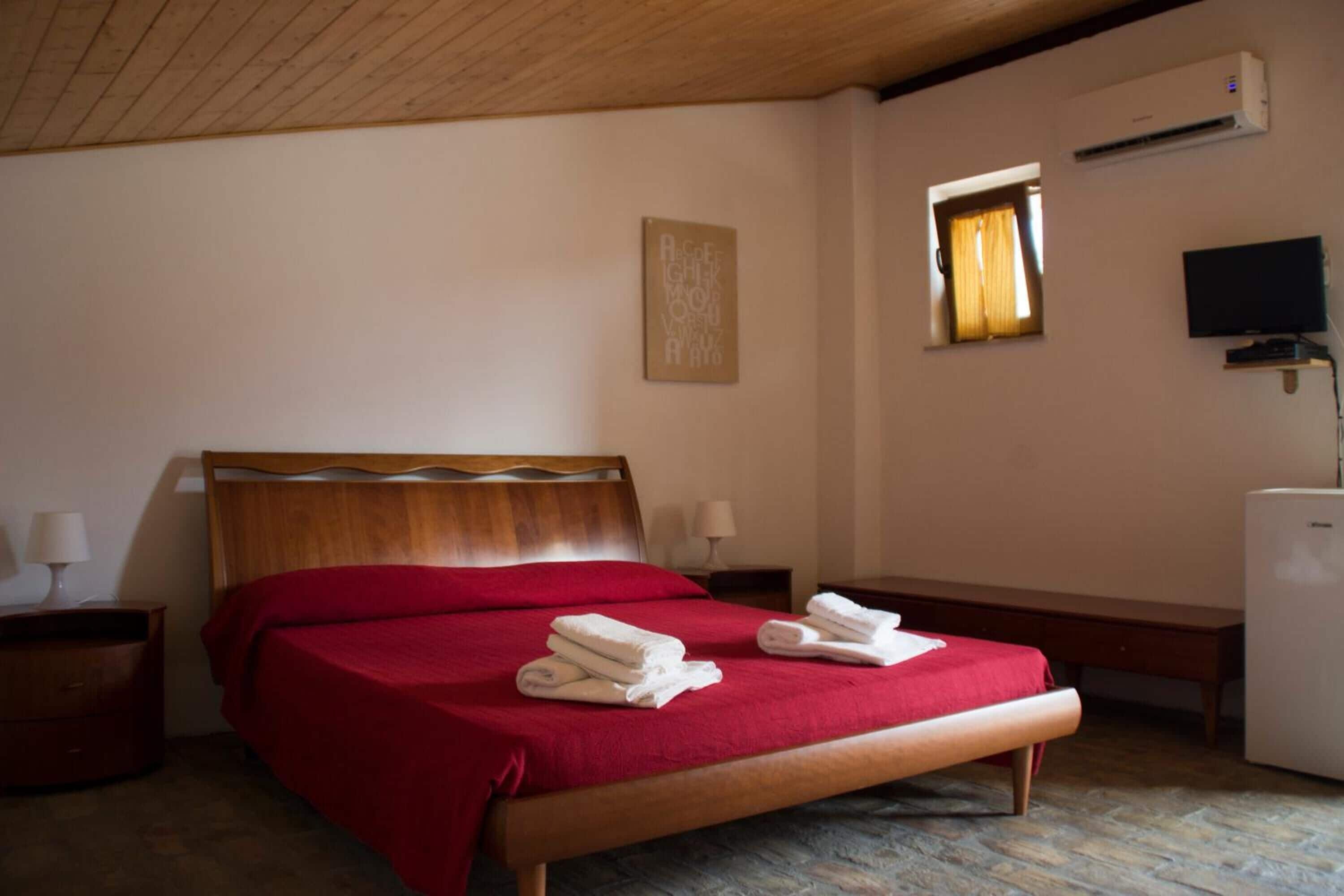 A hotel room with a wooden bed, a small bedside table, a TV, and an air conditioning unit.