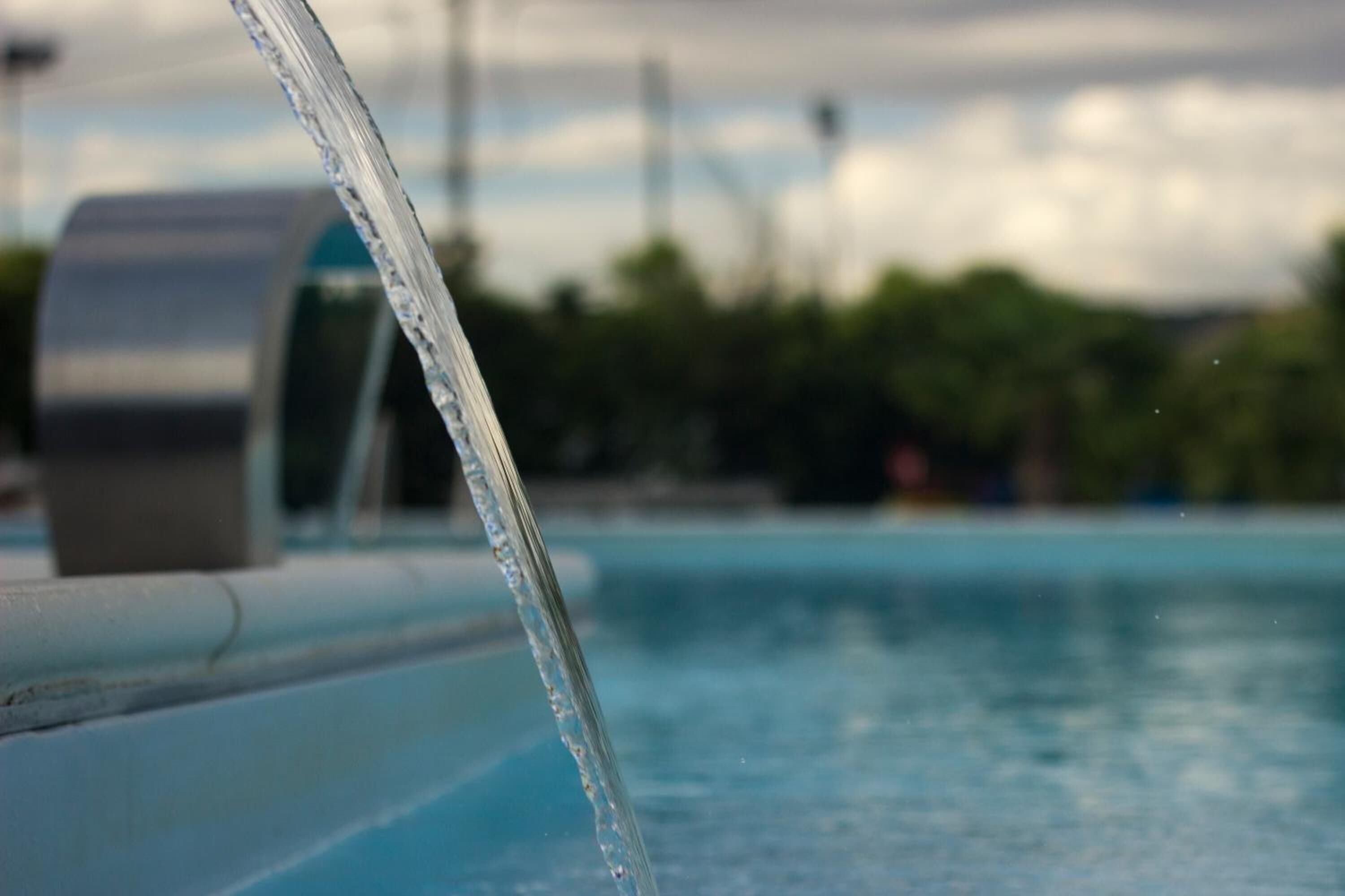 A close-up of a water spout with a blurred background of a swimming pool and greenery.