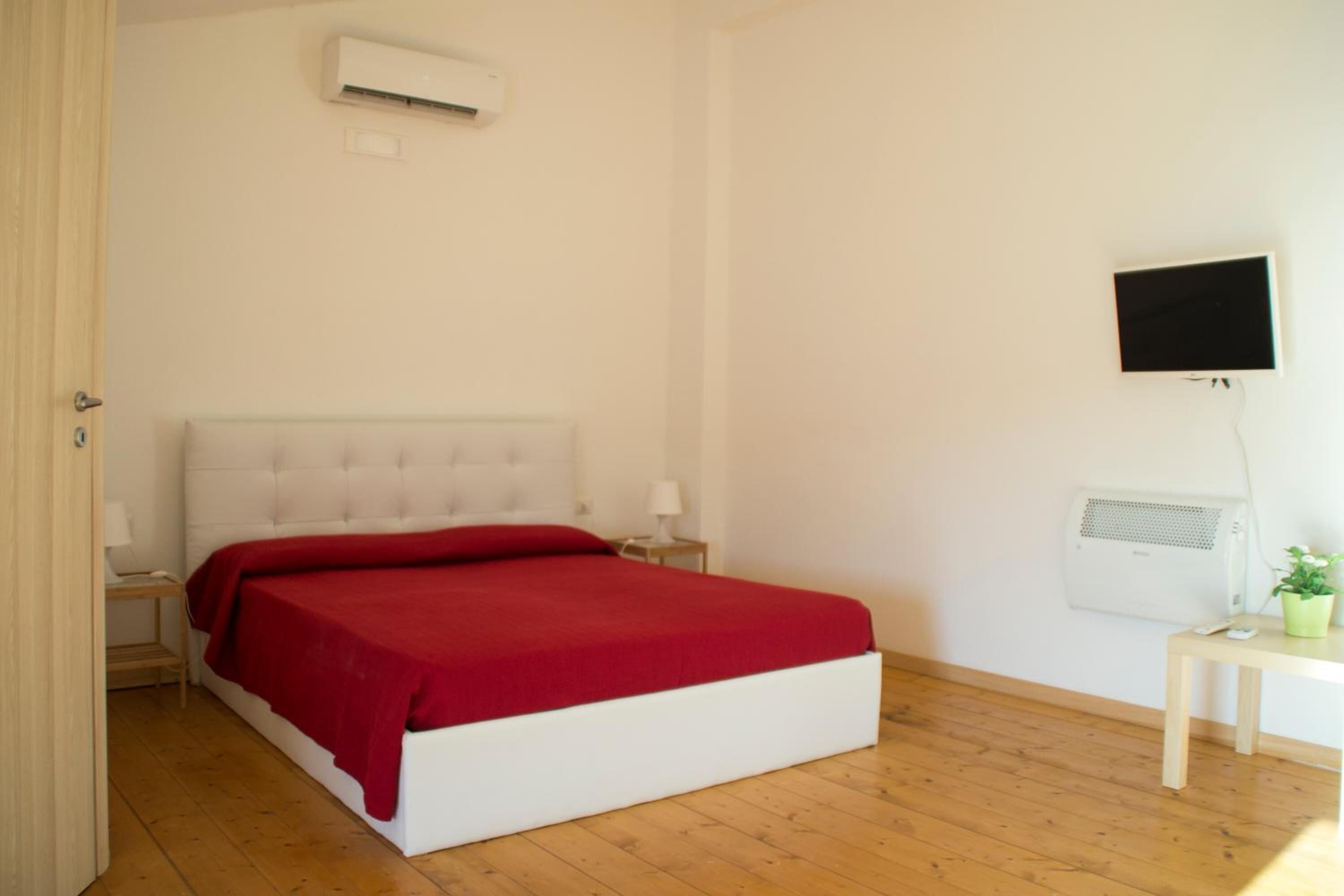 Una camera da letto con un letto bianco, biancheria rossa, un comodino con una lampada, una TV a parete e un condizionatore d'aria.