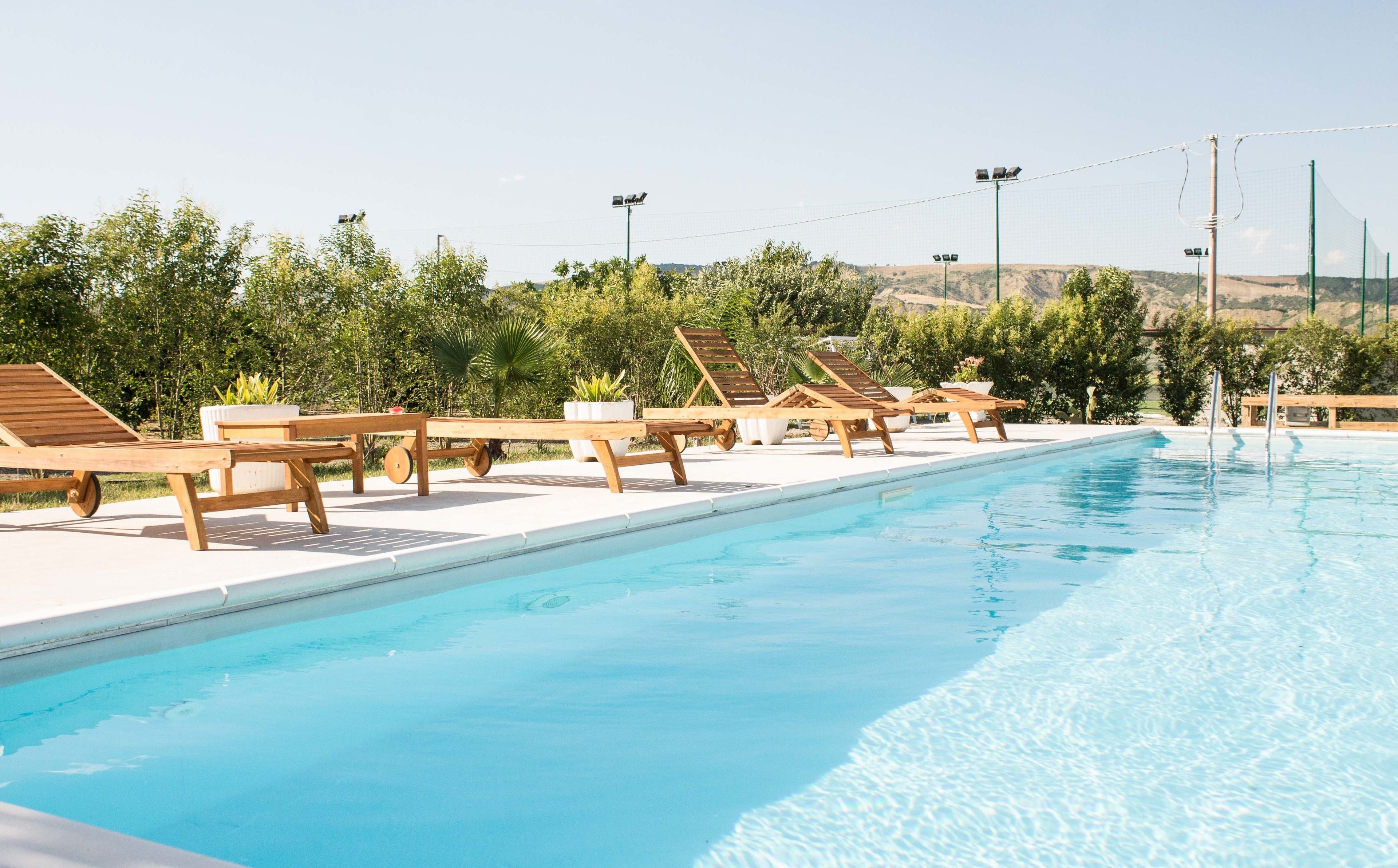 A swimming pool with wooden loungers and potted plants, surrounded by greenery and hills in the background.