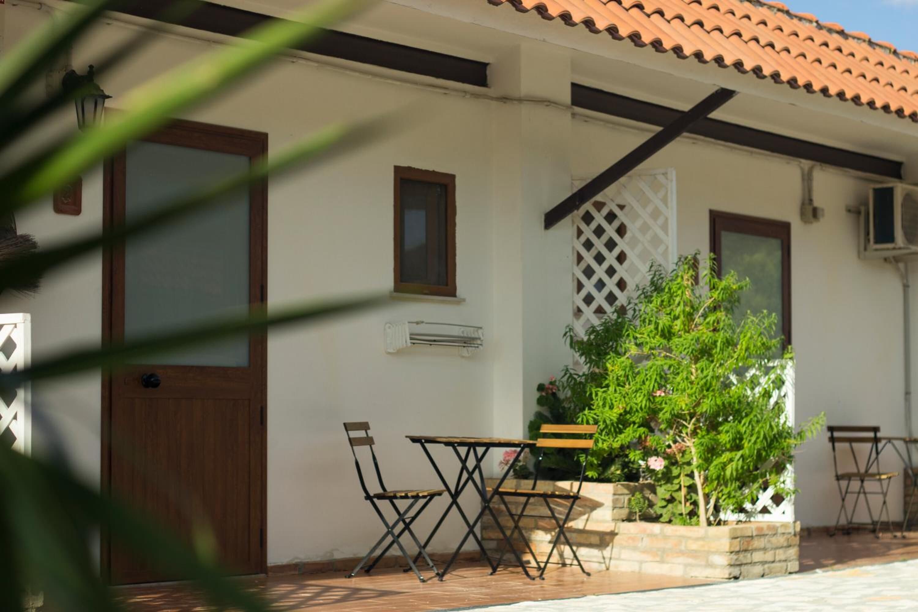 A white building with a tiled roof, a wooden door, and a small table with two chairs. There is a potted plant with green foliage and pink flowers.