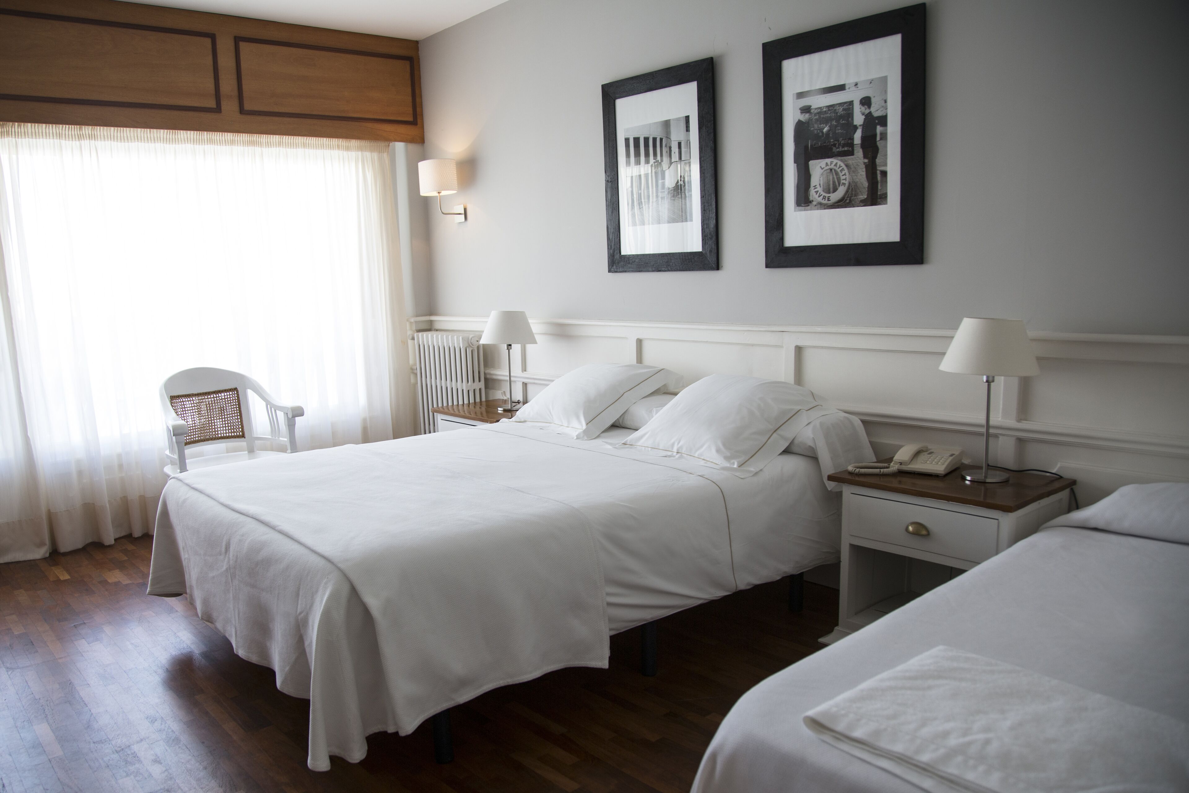 Quarto de hotel com duas camas, uma cadeira, mesa de cabeceira com abajur e quadros nas paredes.