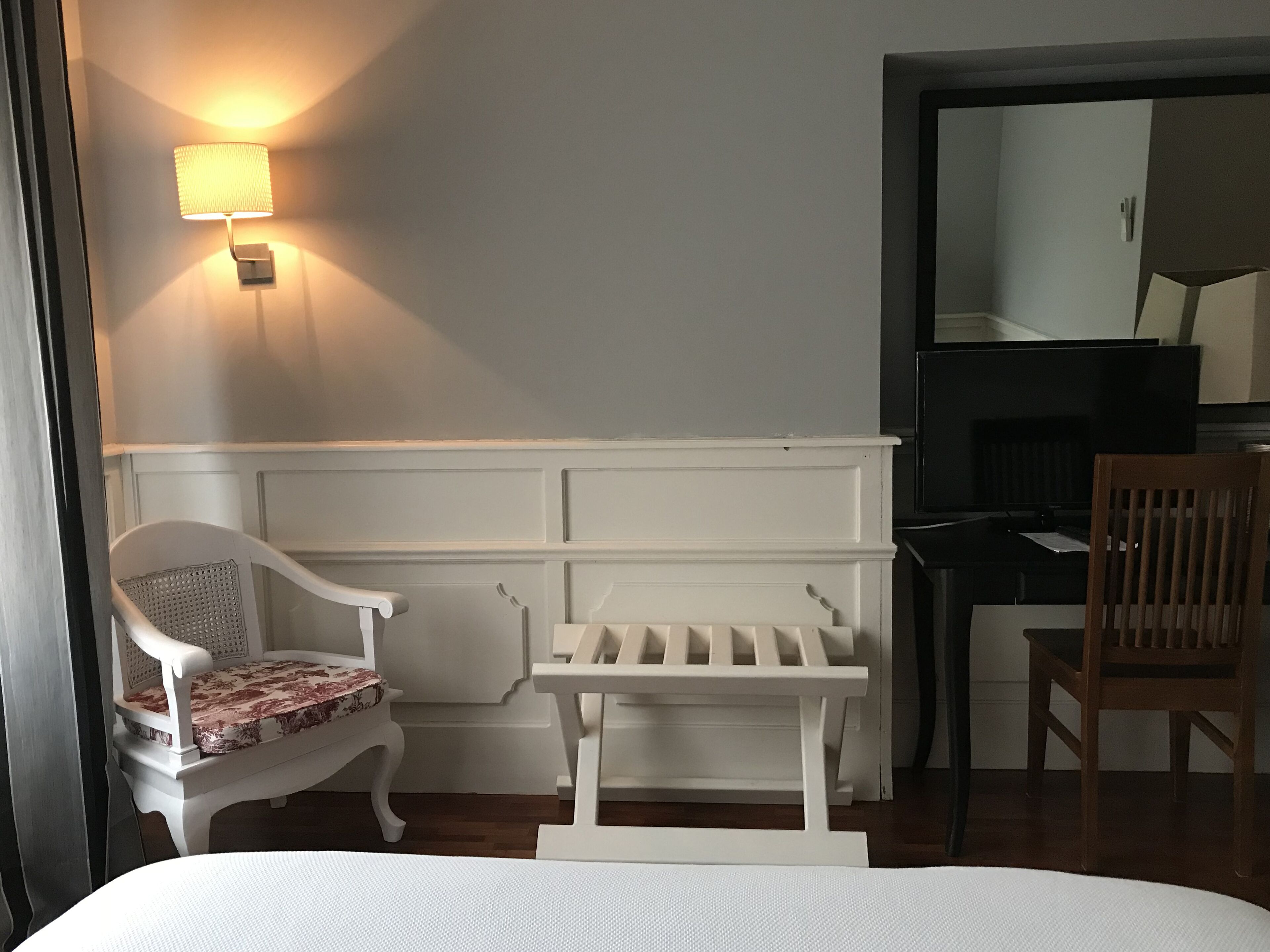 Une chambre d’hôtel avec un lit, un banc blanc, une chaise en bois, un bureau avec un ordinateur et une lampe fixée au mur.