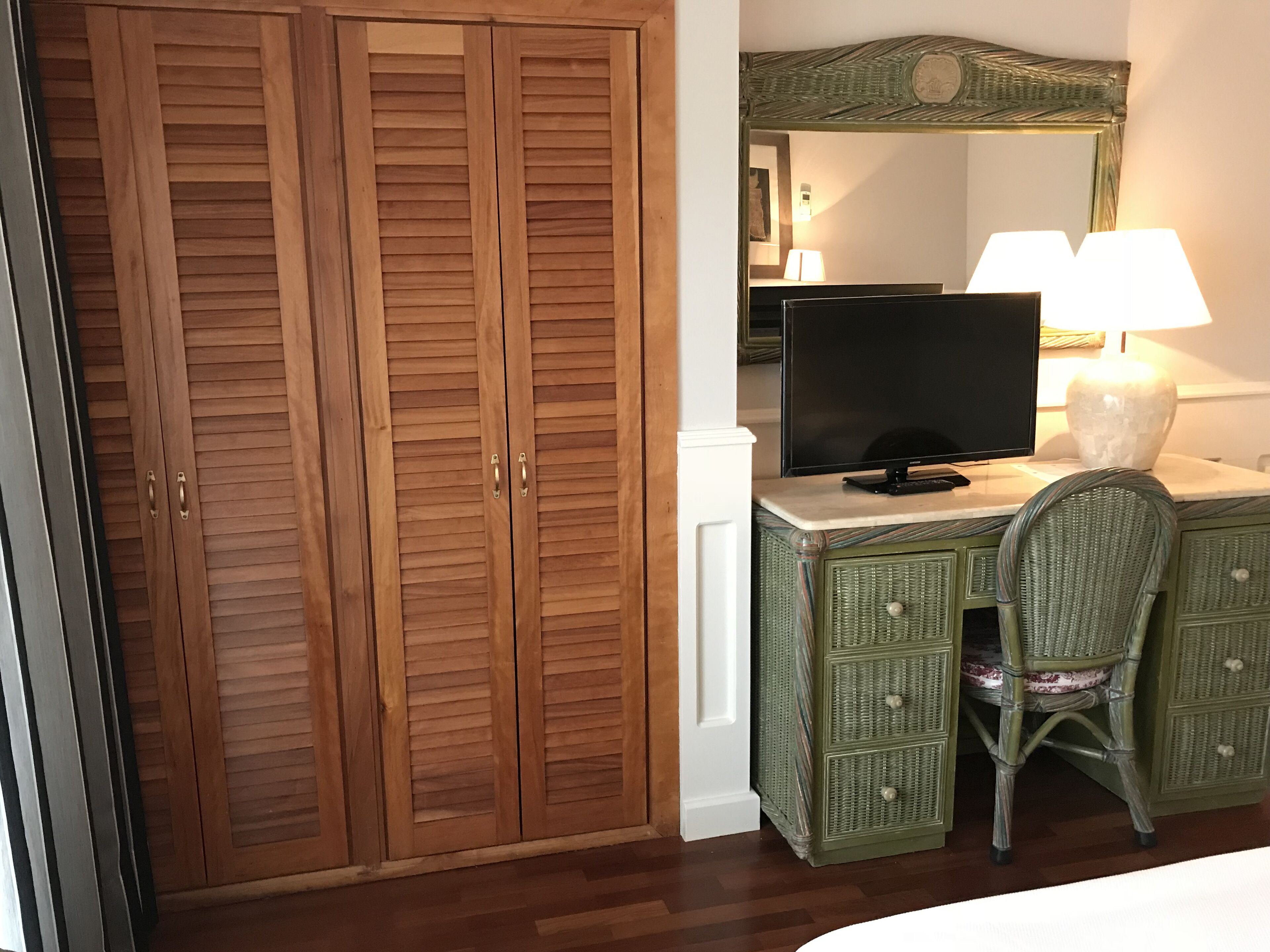 A room with a wooden wardrobe, a television on a marble-topped dresser, a wicker chair, and a lamp.
