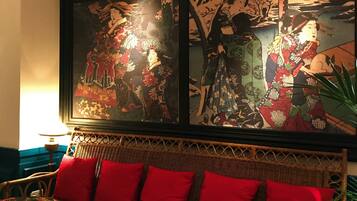 Quarto com uma grande pintura tradicional japonesa na parede, um sofá de vime com almofadas vermelhas e um abajur sobre uma mesinha lateral.