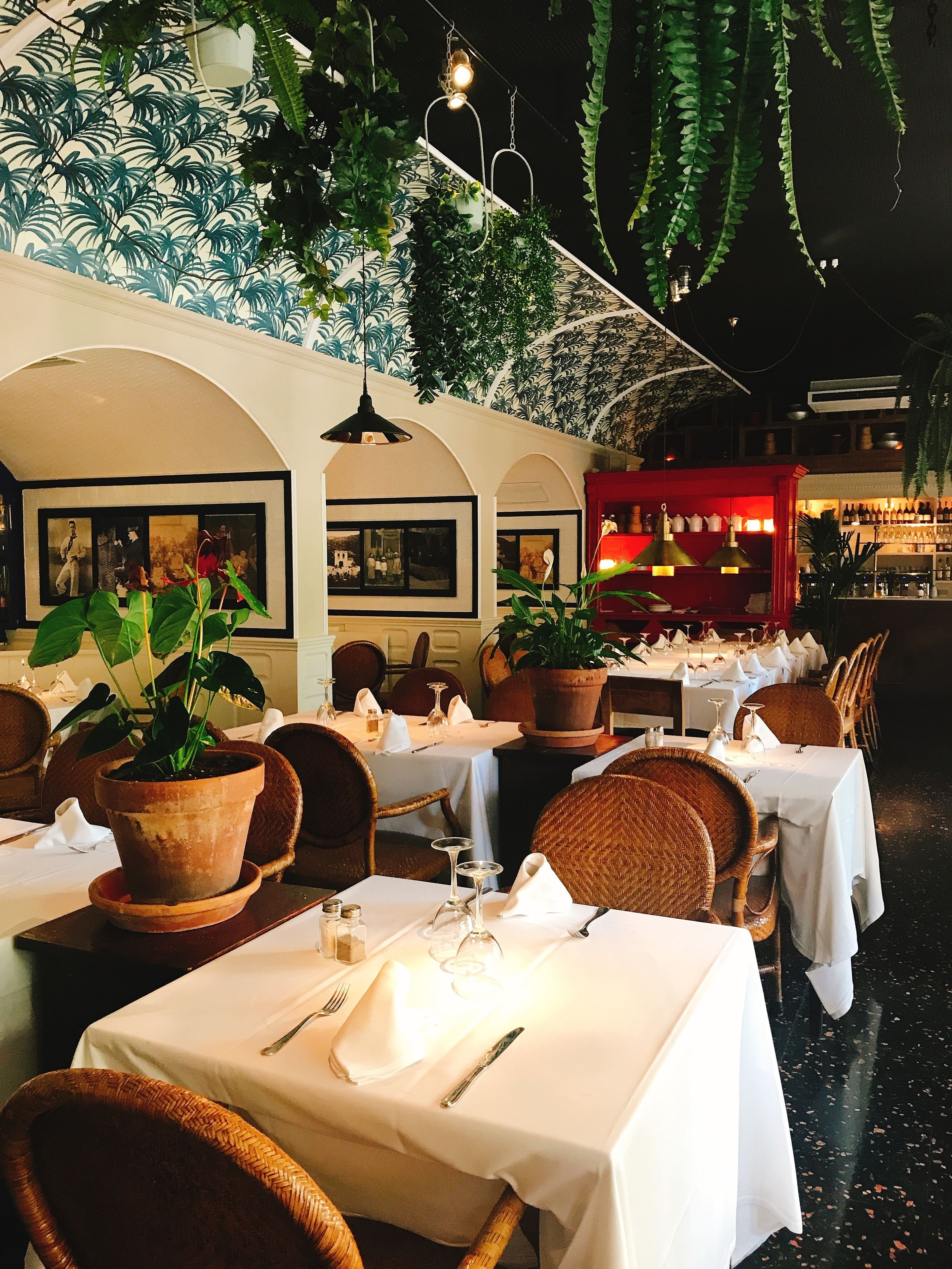 Un comedor interior con mesas preparadas para dos, rodeado de sillas de mimbre y plantas en macetas.