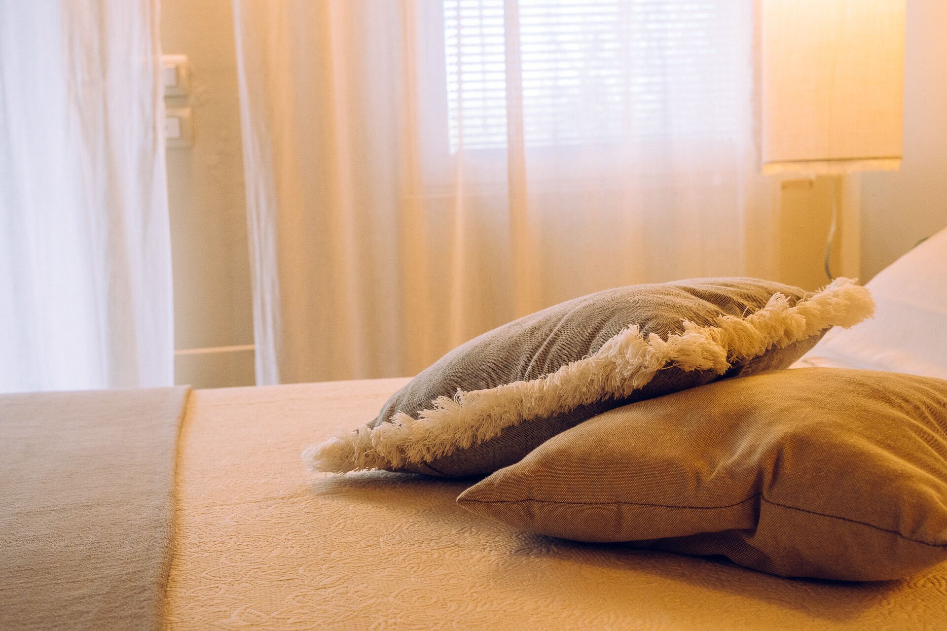 A bed with two pillows, a lamp, and sheer curtains in the background.