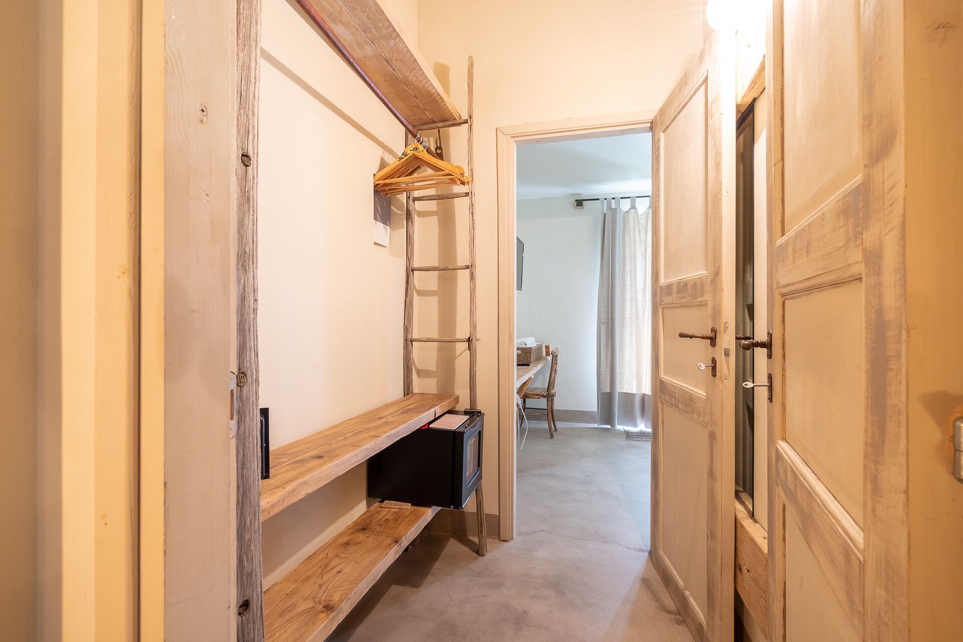 A narrow hallway with wooden shelves, a black cabinet, and a door leading to a room with a chair and a table.