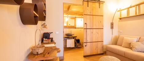 A cozy living room with a wooden bench, a sofa, and a knitted pouf. A sliding wooden door leads to a kitchen area.