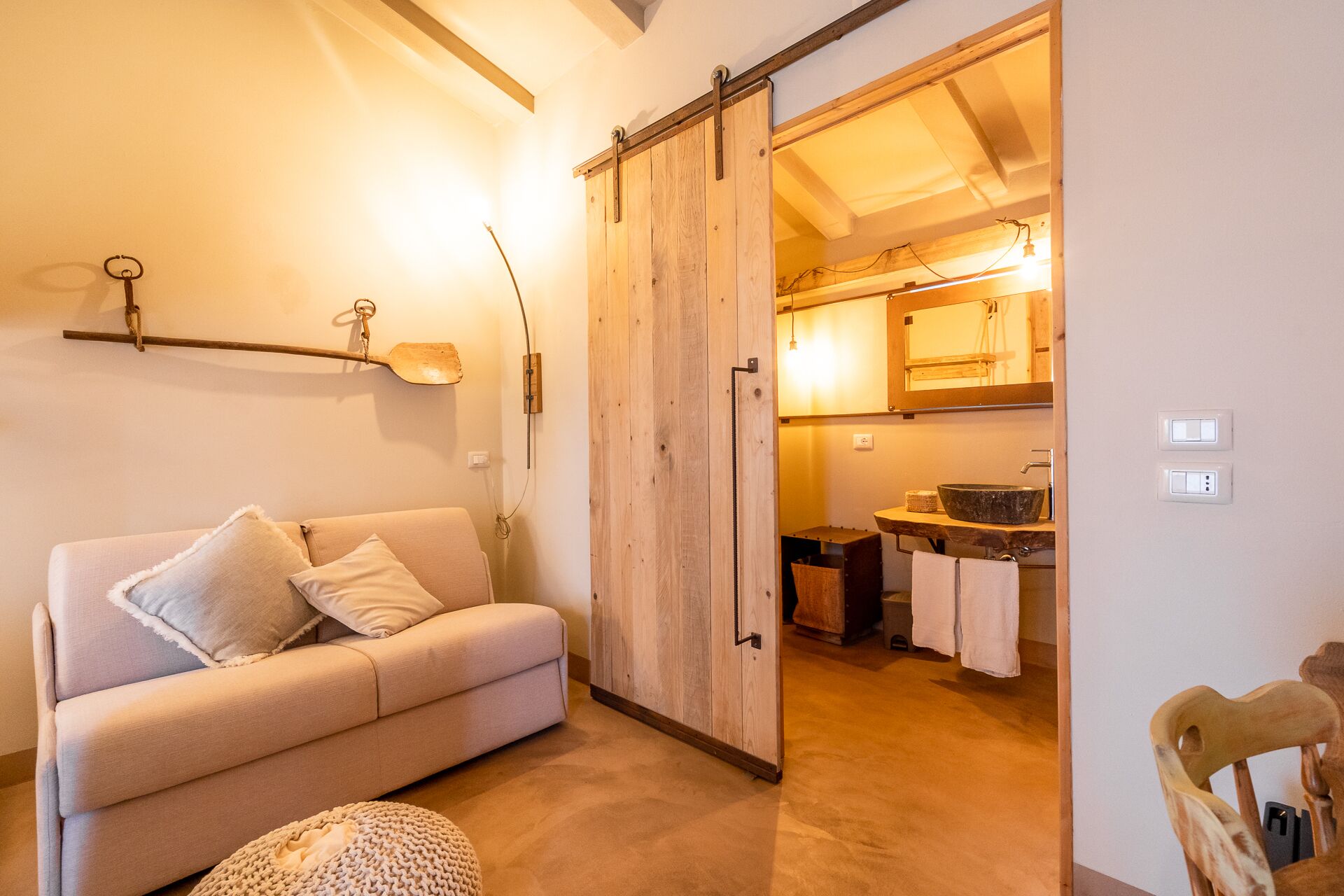 Un salón acogedor con un sofá y reposapiés, y una puerta corredera de madera que da a una cocina con fregadero y encimera.