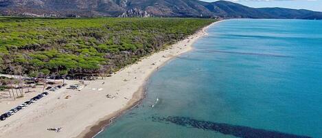 Una playa costera con aguas turquesas cristalinas, una orilla arenosa y una zona boscosa con coches aparcados.