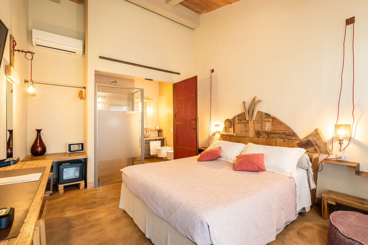 A bedroom with a large bed, a wooden headboard, a desk with a chair, a mirror, and a door leading to the bathroom.