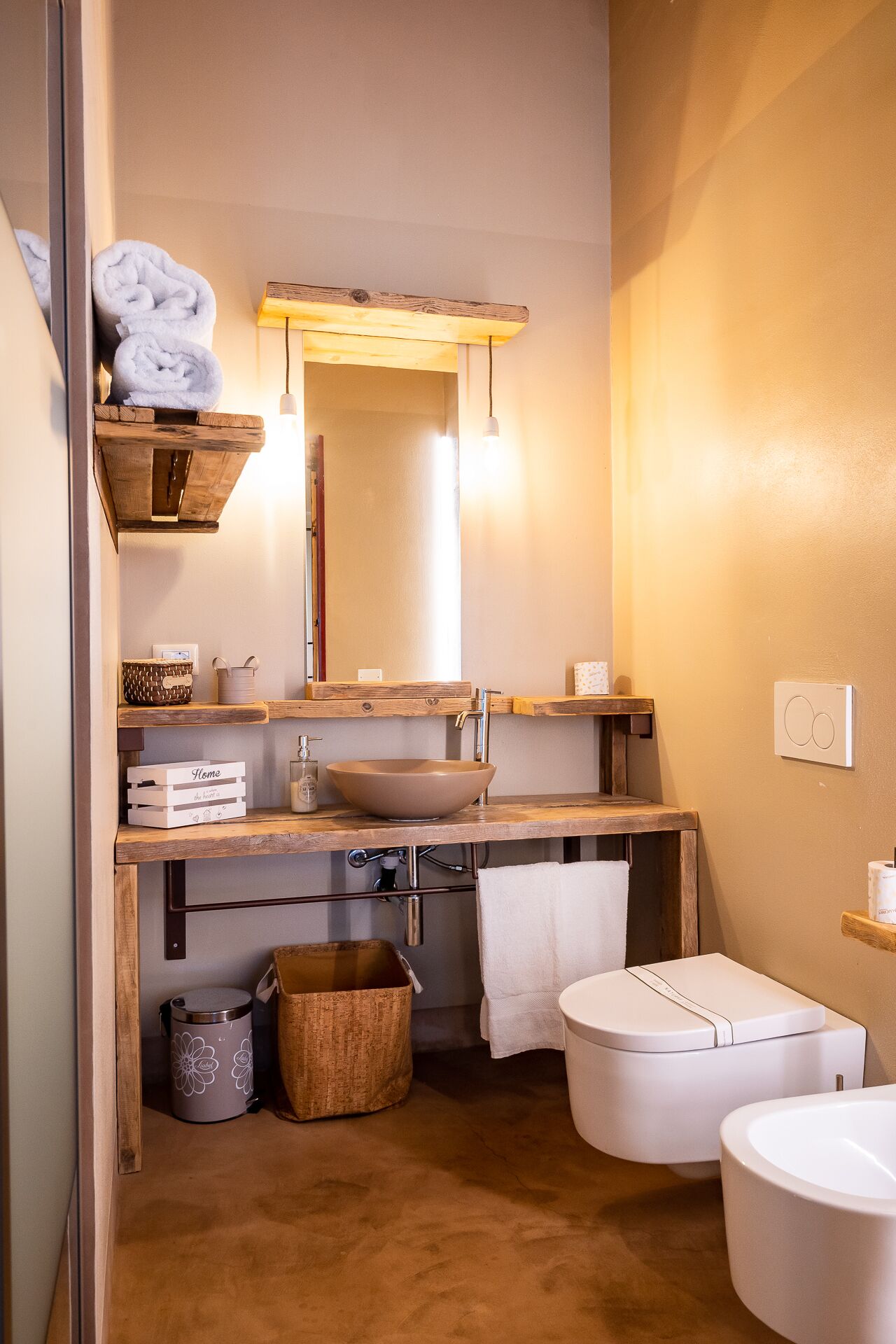 Une petite salle de bain avec un meuble-lavabo en bois, un grand miroir, un lavabo sur colonne, des toilettes et une petite étagère avec des serviettes.