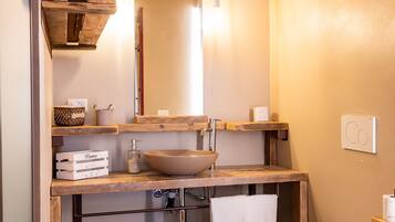 Une petite salle de bain avec un meuble-lavabo en bois, un grand miroir, un lavabo sur colonne, des toilettes et une petite étagère avec des serviettes.