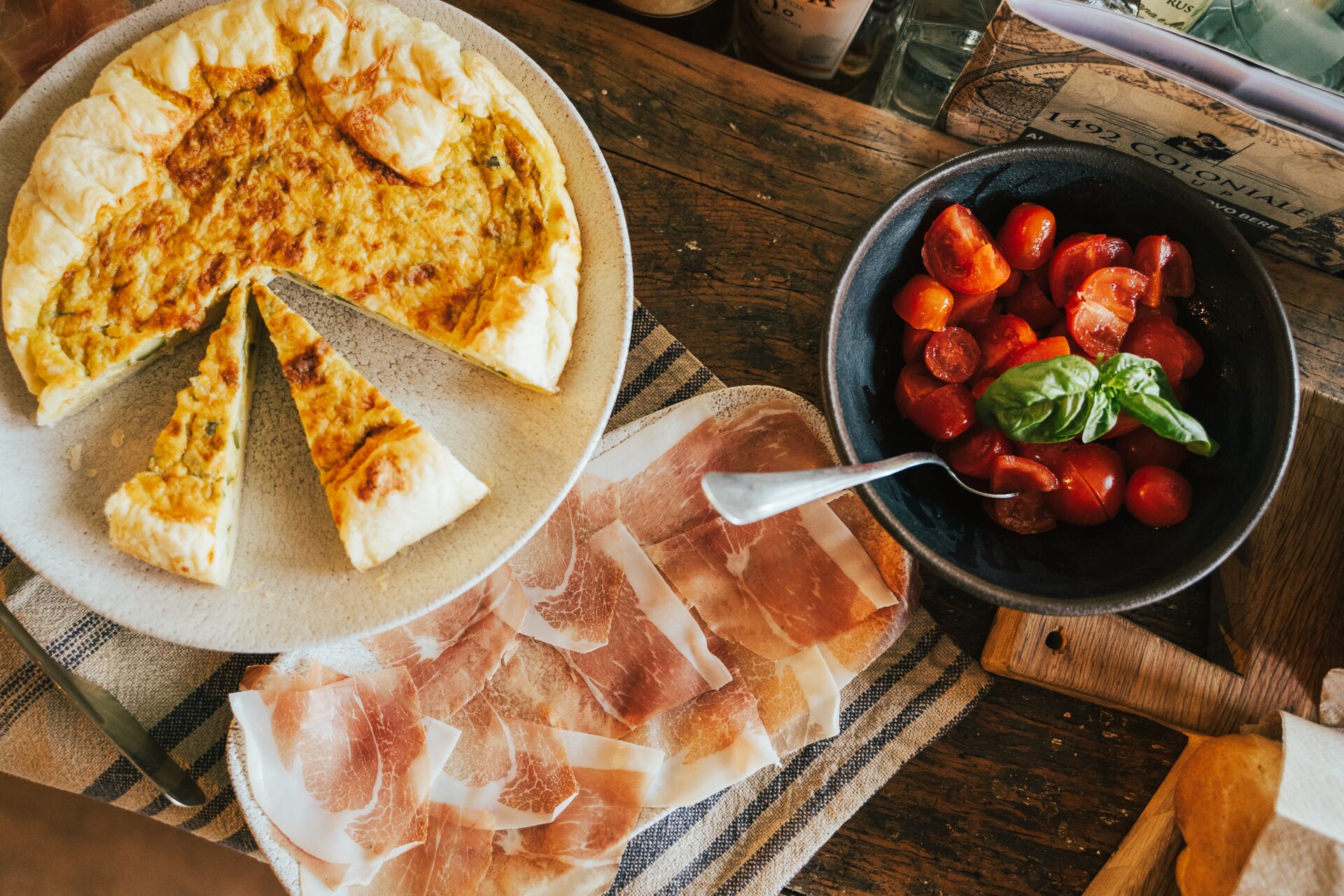 A pizza on a plate, a bowl of tomatoes, and slices of cured meat on a wooden table.