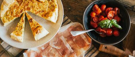 A pizza on a plate, a bowl of tomatoes, and slices of cured meat on a wooden table.