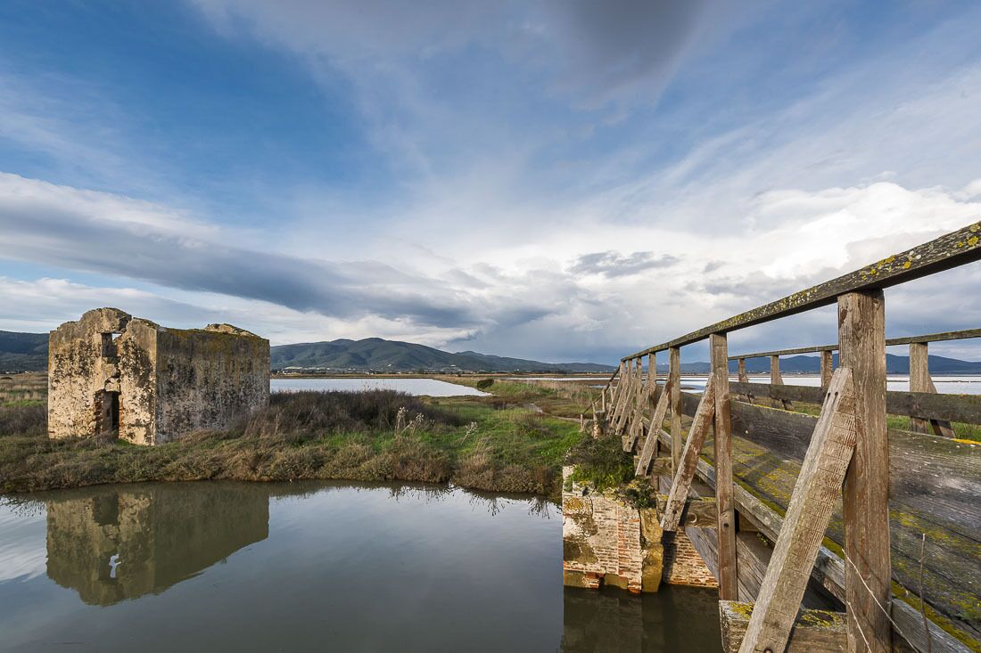 A rustic wooden bridge over a calm body of water, leading to a stone structure with a weathered appearance.