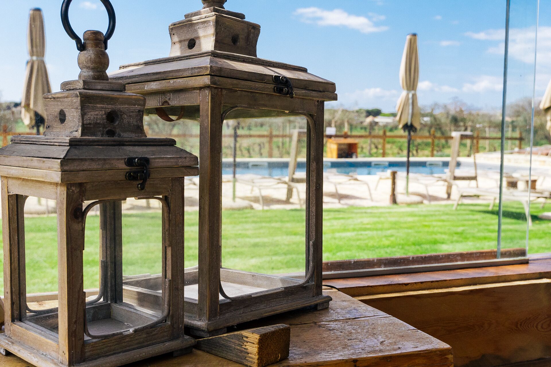 A wooden table with two lanterns and a view of a pool and outdoor seating area.