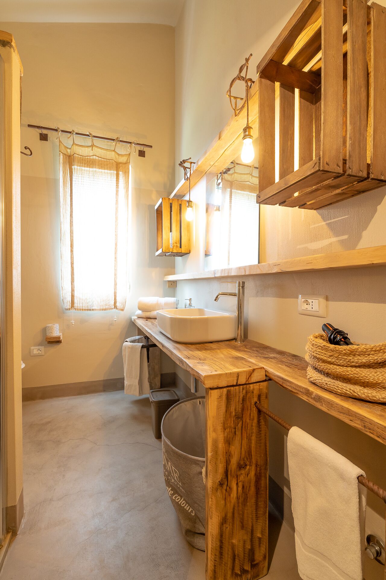 A bathroom with a wooden vanity, a sink, and a mirror. There is a window with curtains, a towel rack, and a trash bin.