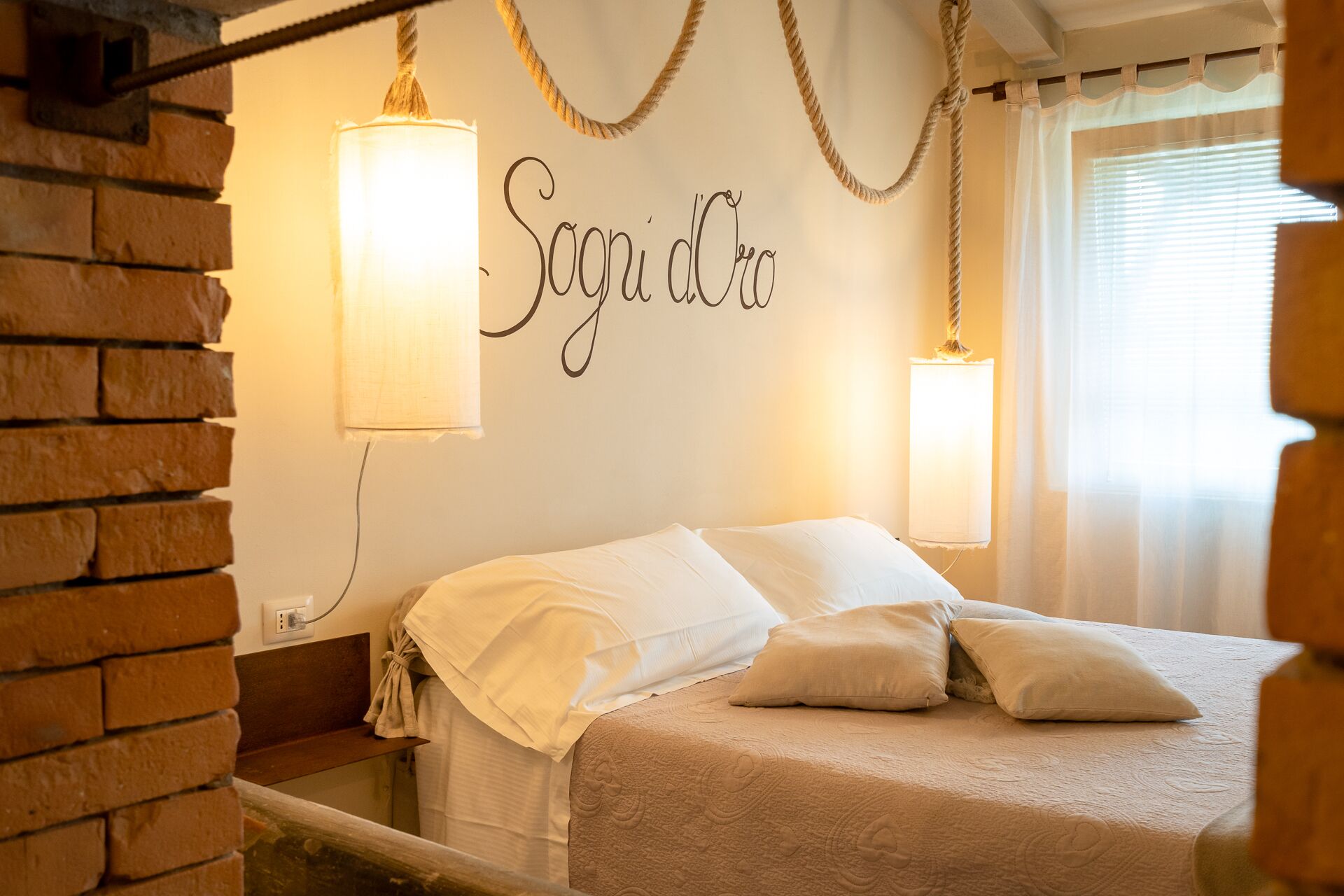 A bedroom with a brick wall, a bed with white bedding, two wall-mounted lamps, and a window with sheer curtains.