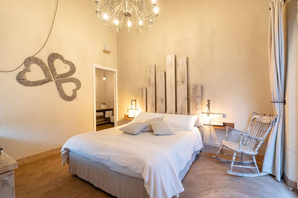 A bedroom with a bed, a rocking chair, a chandelier, and wall art.