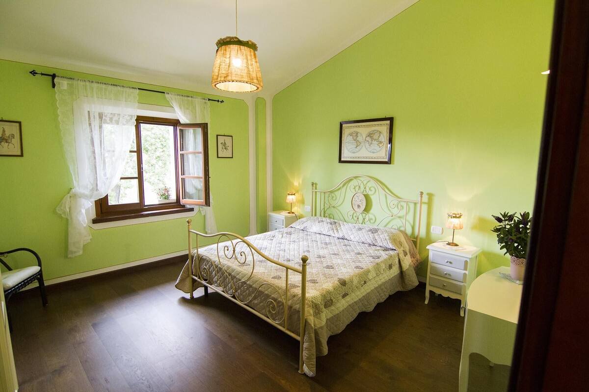Un dormitorio con cama, dos mesitas de noche, ventana con cortinas y una lámpara colgante.