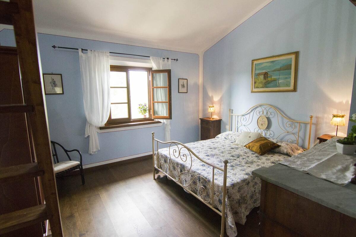 Un dormitorio con cama, ventana con cortinas, mesita de noche con lámpara y un cuadro en la pared.