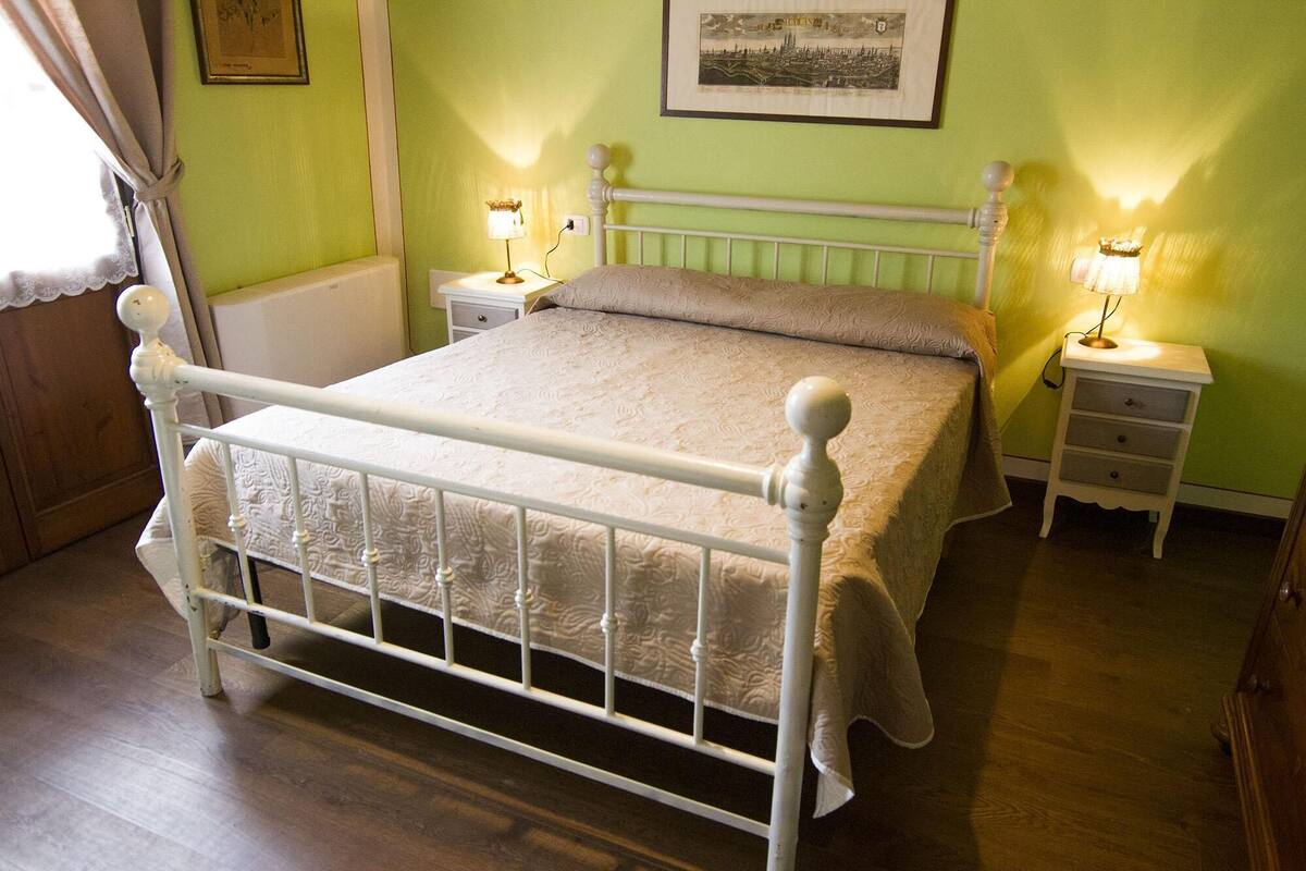Un dormitorio con estructura de cama de metal blanco, colcha color beige, dos mesitas de noche con lámparas y un cuadro en la pared.
